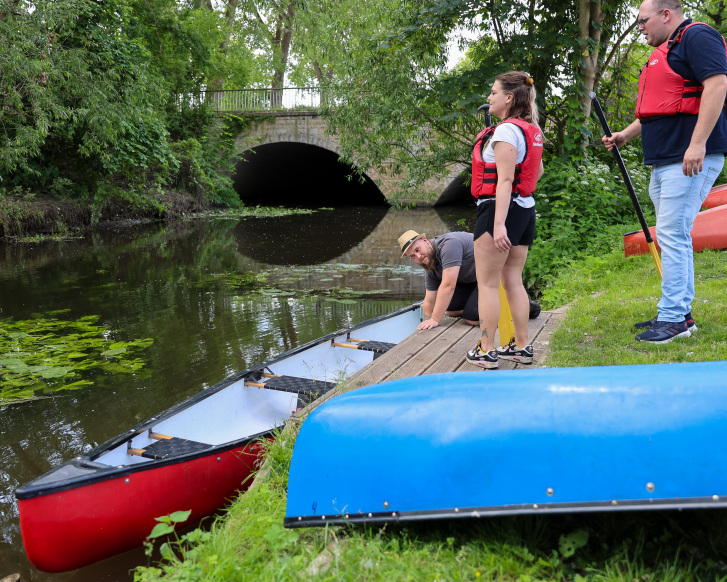 Eine Gruppe von Leuten, in Schwimmwesten, am Ufer eines Flusses mit Kanus und Rudern, in Brückennähe.
