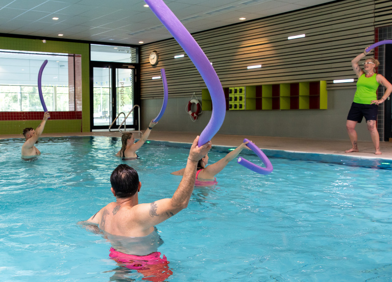 Menschen mit Schwimmnudeln in den Händen betätigen sich sportlich in einem Schwimmbecken. Eine Anleiterin macht am Beckenrand die Übungen vor.