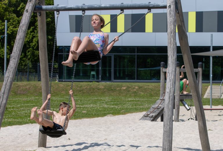 Zwei Kinder in Badekleidung schaukeln auf einem Spielplatz. Der Untergrund ist Sand, im Hintergrund sieht man ein sehr kleines Klettergerüst aus Holz.