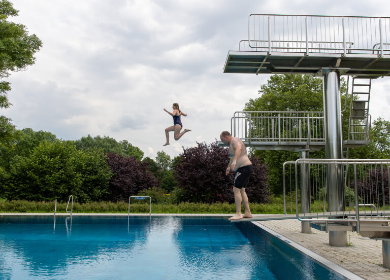 Ein Aussenschwimmbecken mit Drei verschieden hohen Sprungtürmen. Ein Mann möchte gerade vom niedrigsten Brett springen. Ein Mädchen ist soeben vom mittlerem Brett abgesprungen.