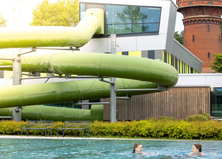 Eine grüne Röhrenrutsche kringelt sich über einem Aussenbecken in dem zwei Mädchen schwimmen.