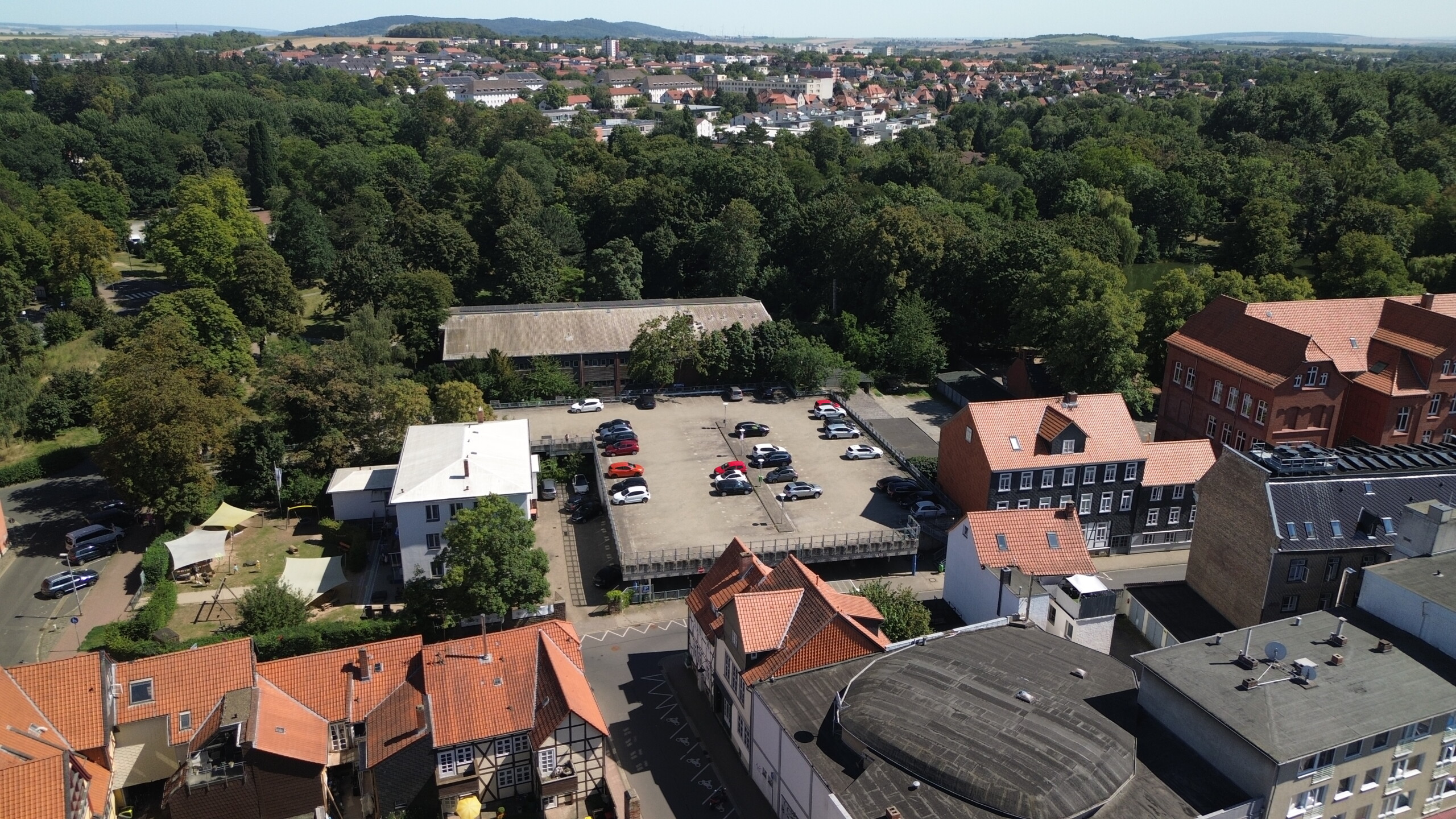 Eine große, sonnige Parkplatzfläche von oben, zwischen Häusern und Bäumen, in der Ferne sieht man eine Stadt.