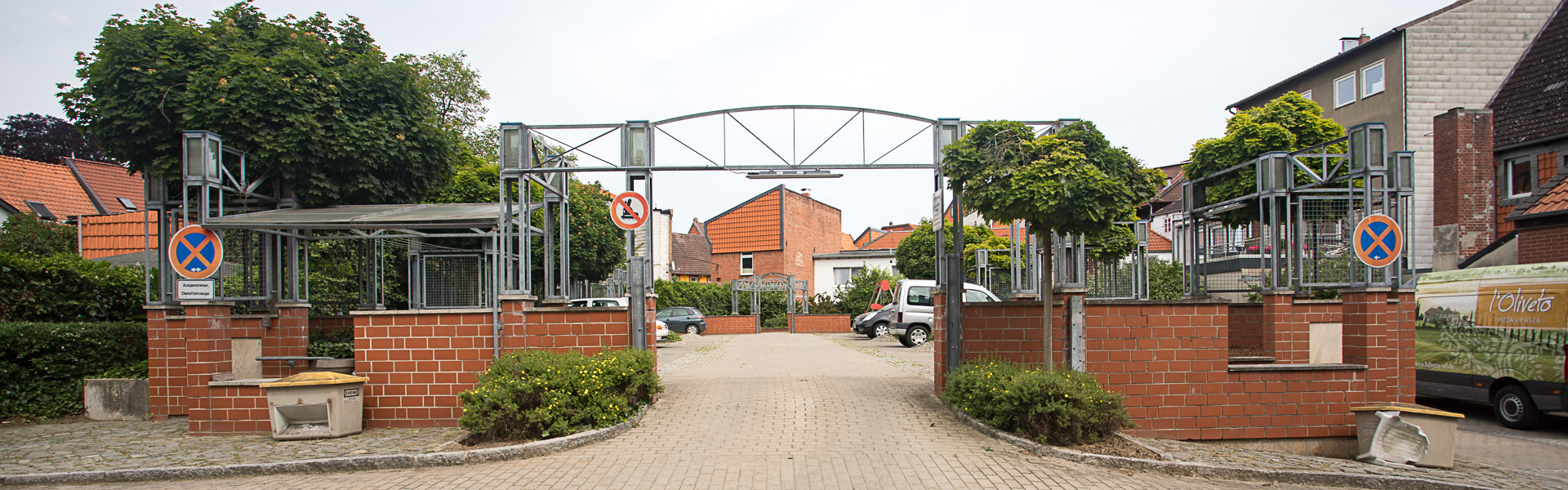 Ein mit roten Klinkersteinen halbhoch eingezäunter Parkplatzbereich mit einem stählernen Torbogen zur Einfahrt.